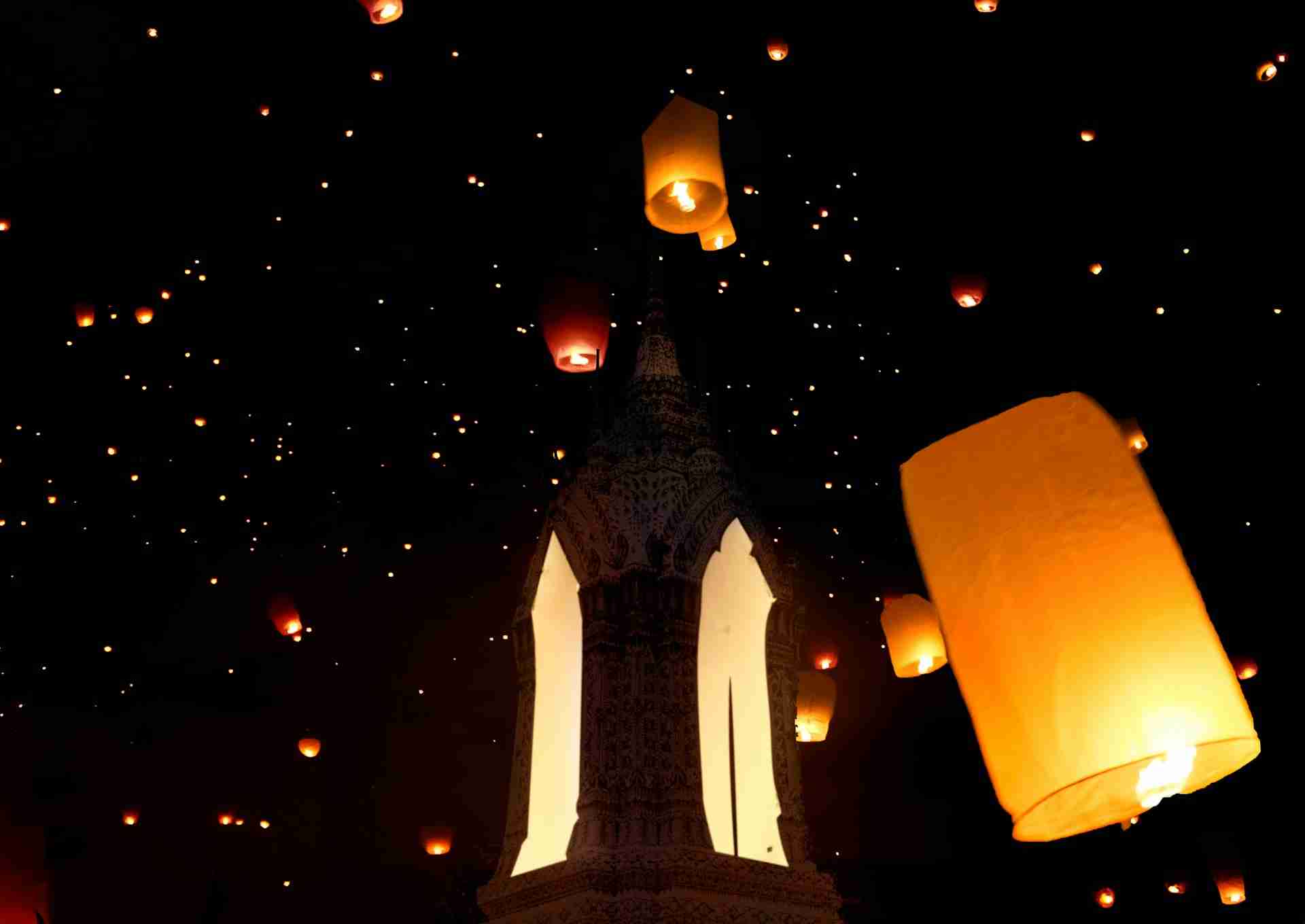 The image shows a night scene of glowing sky lanterns floating above an illuminated, ornate temple or monument, creating a magical and festive atmosphere.